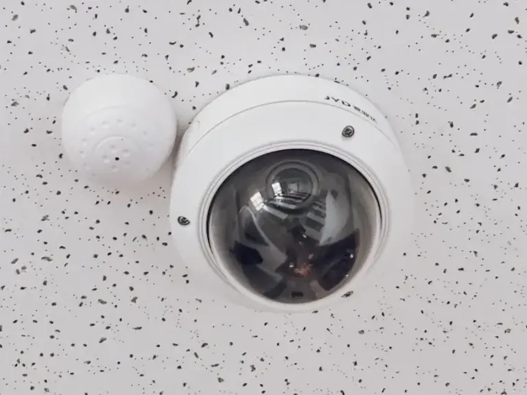 White dome security camera and round sensor mounted side by side on a speckled ceiling tile