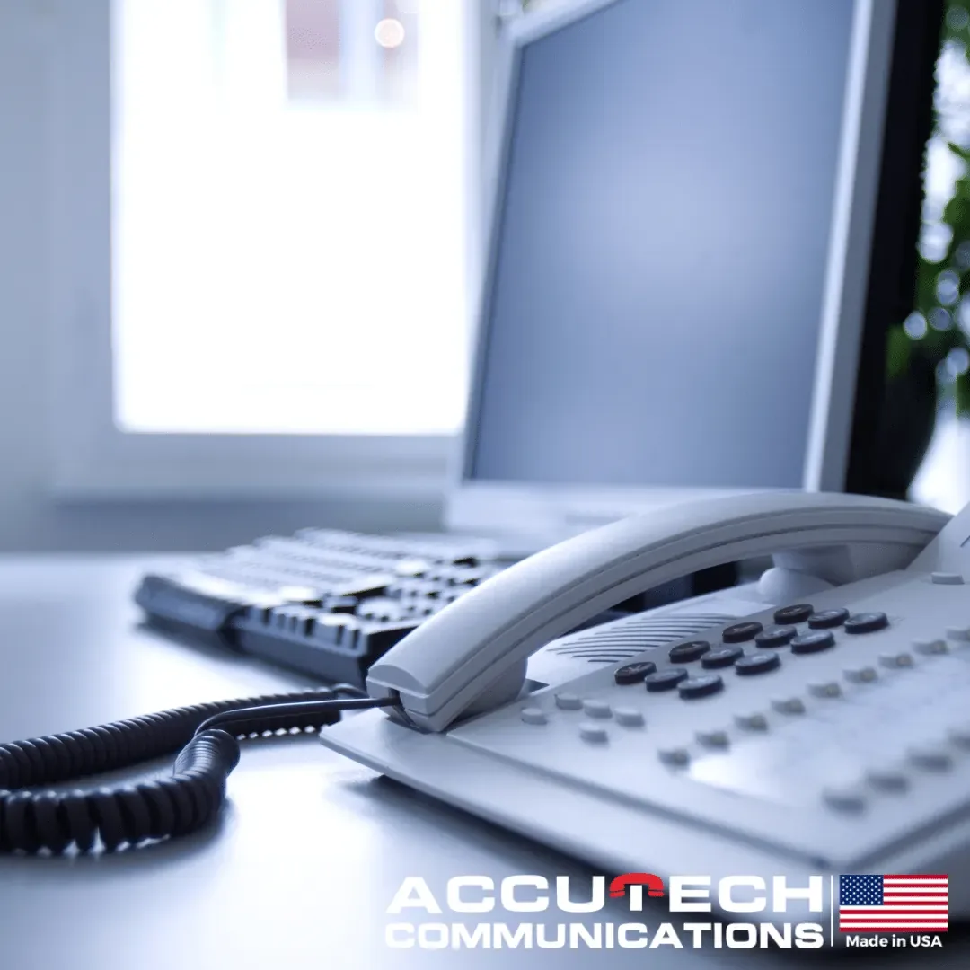Gray corded desk phone beside a keyboard and monitor on a bright office desk