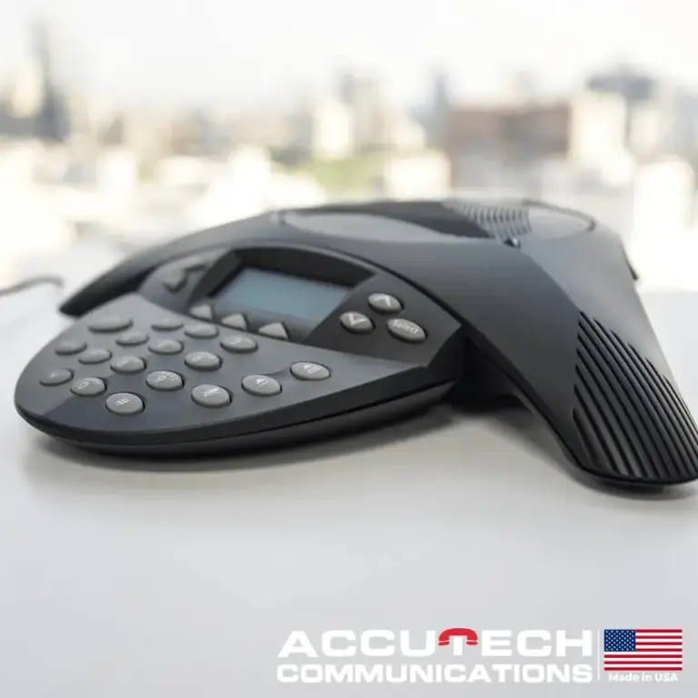 Black conference speakerphone with display and keypad on a white table with a blurred city view
