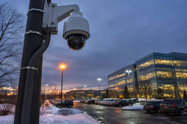 Pole-mounted pan-tilt-zoom security camera over a snowy office park parking lot at dusk