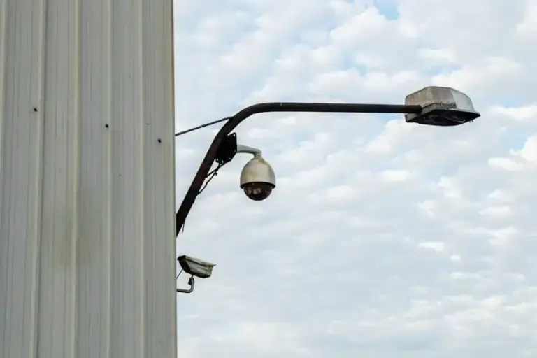 Dome camera and older box camera mounted on a pole arm with a light against a cloudy sky