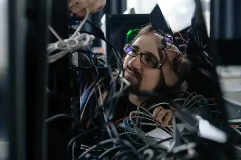Man in glasses peering through a tangle of gray cables behind an open server rack