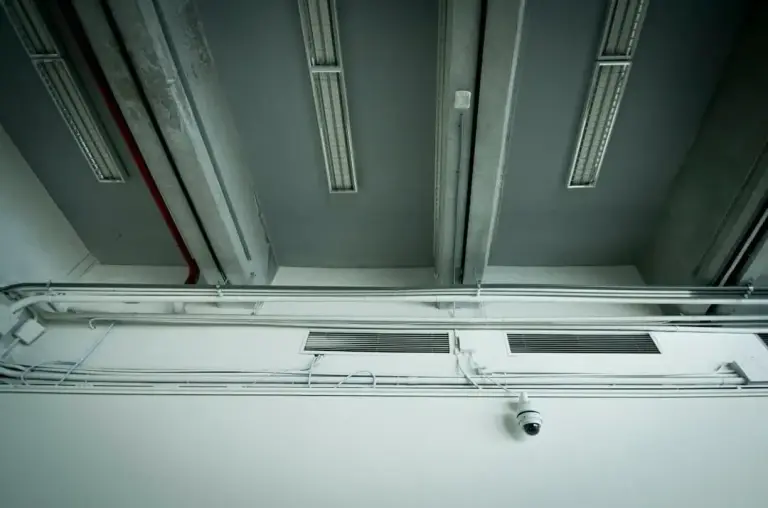 Small dome camera mounted on a white wall below conduit runs and an exposed ceiling