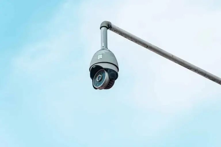 Gray dome security camera hanging from a metal pole arm against a pale sky